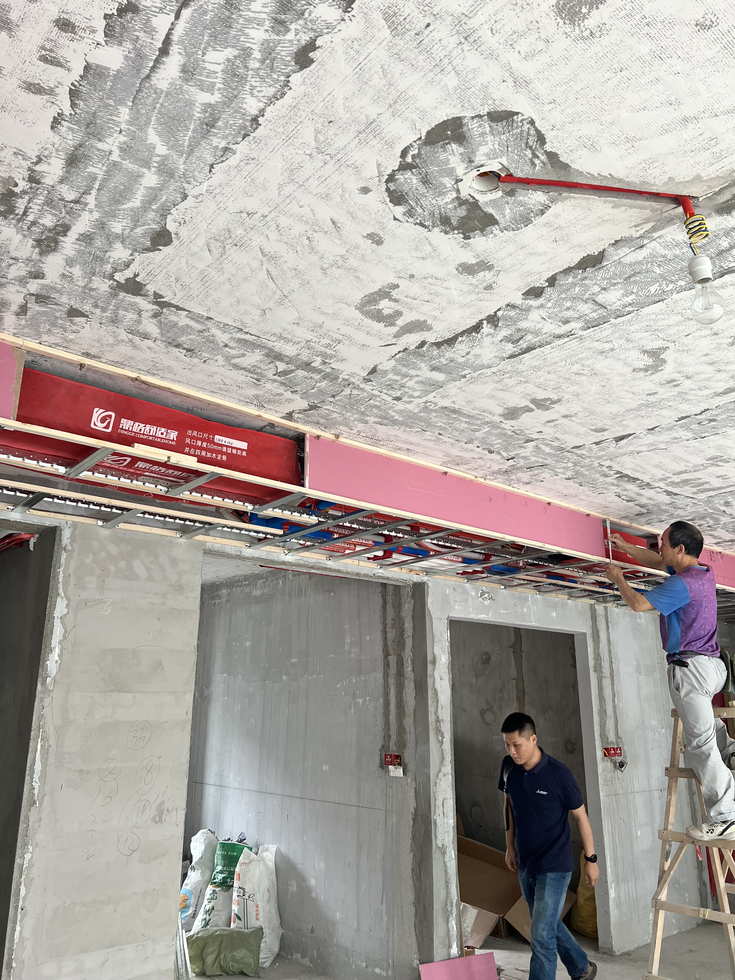 毛坯室內吊頂下方已就位的中央空調室內機，機身套有紅色品牌保護罩，兩名工人在梯子上進行吊頂與機器定位作業